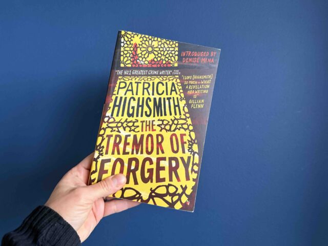 Cover of the Virago Modern Classics edition with brown letters against a yellow background, held by my hand against a blue background.