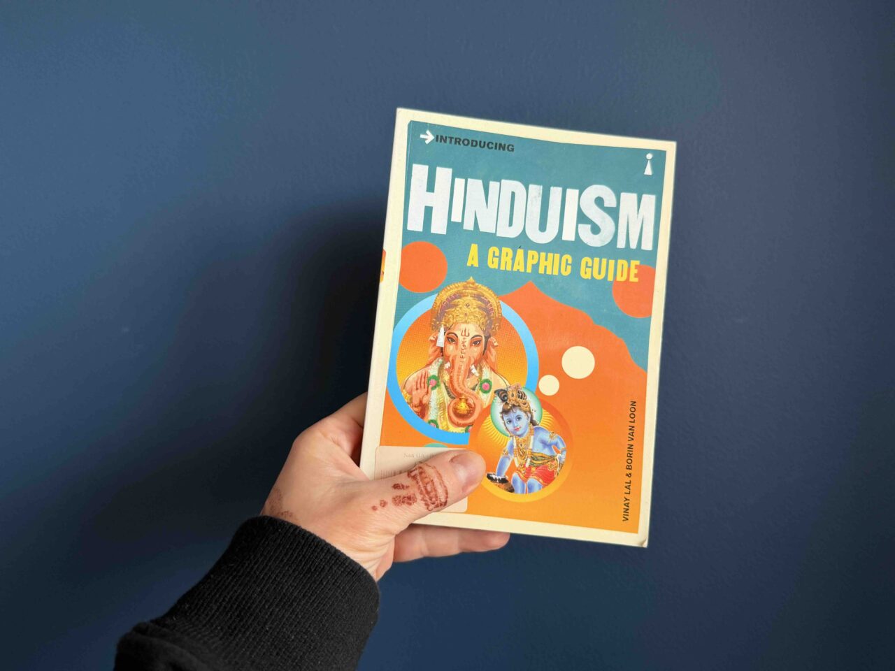 My hand (with henna tattoos, holding a paperback copy of the book, with a drawing of Ganesha, the elephant god, against a navy background.
