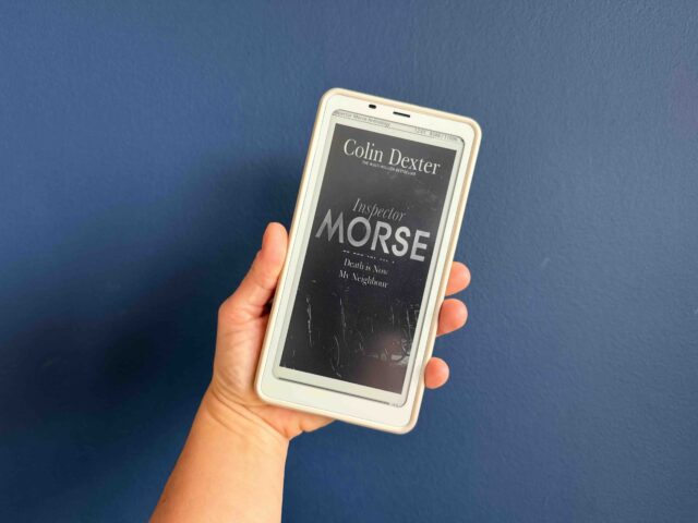 Ebook cover of Death is Now My Neighbour held by my hand against a navy background